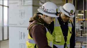 two trades workers in safety helmets and vests looking down